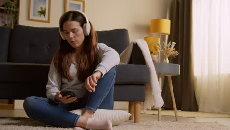 Woman-Wearing-Wireless-Headphones-Sitting-On-Floor-At-Home-Streaming-Music-From-Mobile-Phone-1